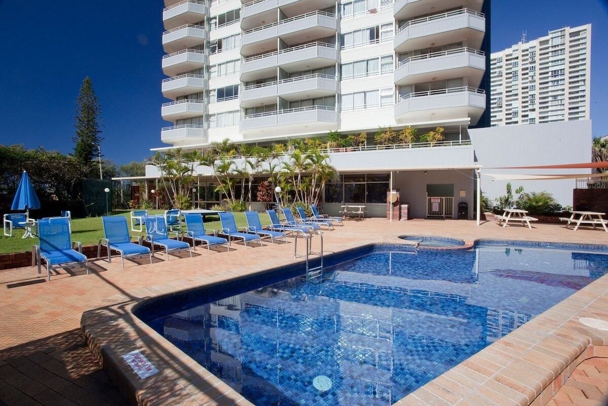 A swimming pool with lounge chairs, a building with multiple balconies, and a clear blue sky.