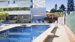 Une piscine entourée d’un bâtiment doté de balcons et de palmiers, avec des chaises longues bleues et des parasols.