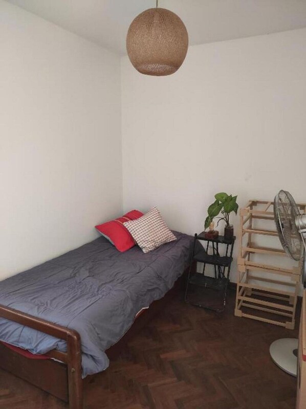 Una habitación pequeña con cama individual, suelo de madera, colcha gris, almohada roja y blanca, lámpara colgante, una mesa pequeña con una planta y un ventilador de pie.