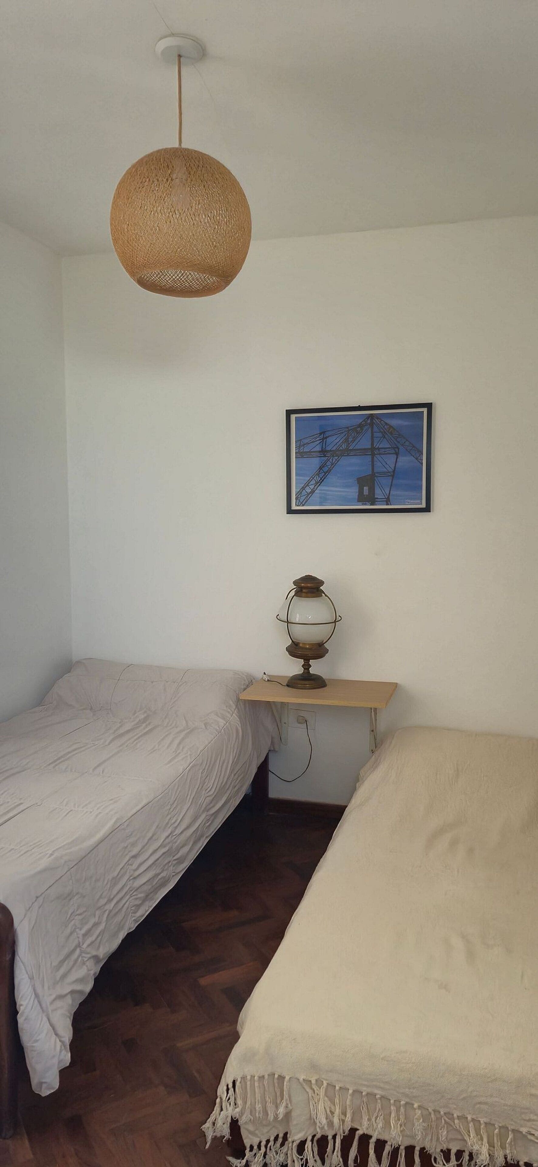 Une petite chambre simple, avec deux lits simples, une petite table avec une lampe et un tableau encadré au mur.