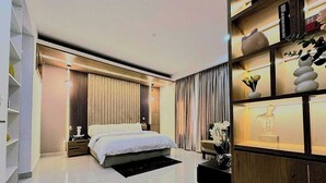 A modern bedroom with a large bed, a bedside table, a nightstand, a wooden cabinet with decorative items, and a bookshelf. - Damido's Apartment (Abuja)