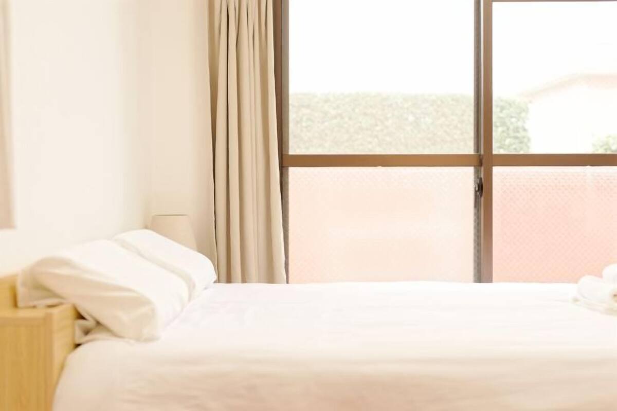 A hotel room with a bed, a nightstand, a window with curtains, and a view of greenery outside.