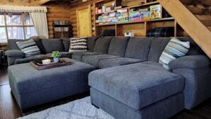 L’intérieur d’une cabane en rondins, avec un grand canapé d’angle, des poutres en bois et une bibliothèque.