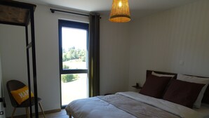 A bedroom with a bed, a chair with a 'HAPPY' pillow, a hanging light fixture, and a view through a window.
