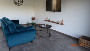 A modern living room with a teal sofa, a round coffee table, a wall-mounted TV, and a minimalist shelf with books and a plant.