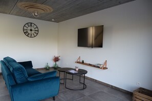 Een moderne woonkamer met een blauwgroene bank, een ronde salontafel, een aan de muur bevestigde televisie, en een minimalistische plank met boeken en een plant.