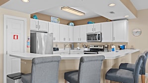 Una cocina moderna con armarios blancos, un refrigerador de acero inoxidable, un microondas y un fregadero. Hay tres taburetes grises y un bote de basura.