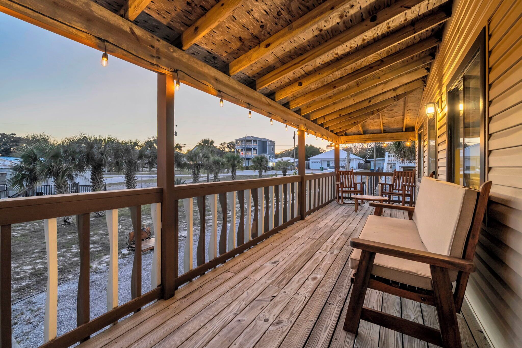 A wooden deck with a railing, a cushioned bench, and a view of palm trees and buildings.