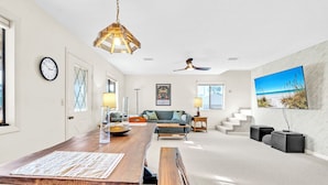 A spacious living area with a wooden dining table, a sofa, a TV mounted on the wall, and a staircase leading to another level.
