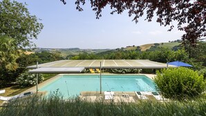 Ein Schwimmbecken mit Sonnenschirm, umgeben von Grünflächen und Hügeln im Hintergrund.