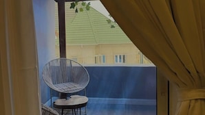 A balcony with a wicker chair and a small table, a view of a building with a green roof, and a curtain partially drawn. - The Pines Court Apartment (Gwagwalada)