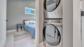 Une chambre à coucher comprenant un lit, une petite table de chevet, un lave-linge fixé au mur et une fenêtre donnant sur un paysage verdoyant.