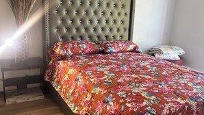 Camera da letto con testiera capitonata, copriletto floreale e comodino con un vaso di piante essiccate.