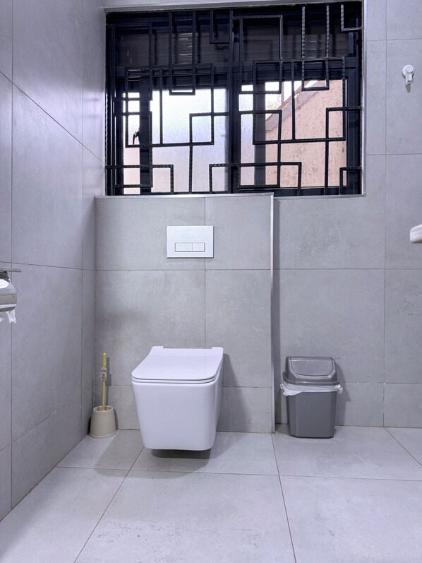 A small, clean bathroom with a toilet, a trash bin, and a window with a black metal grille. - Jay Jay Homes (Kampala)