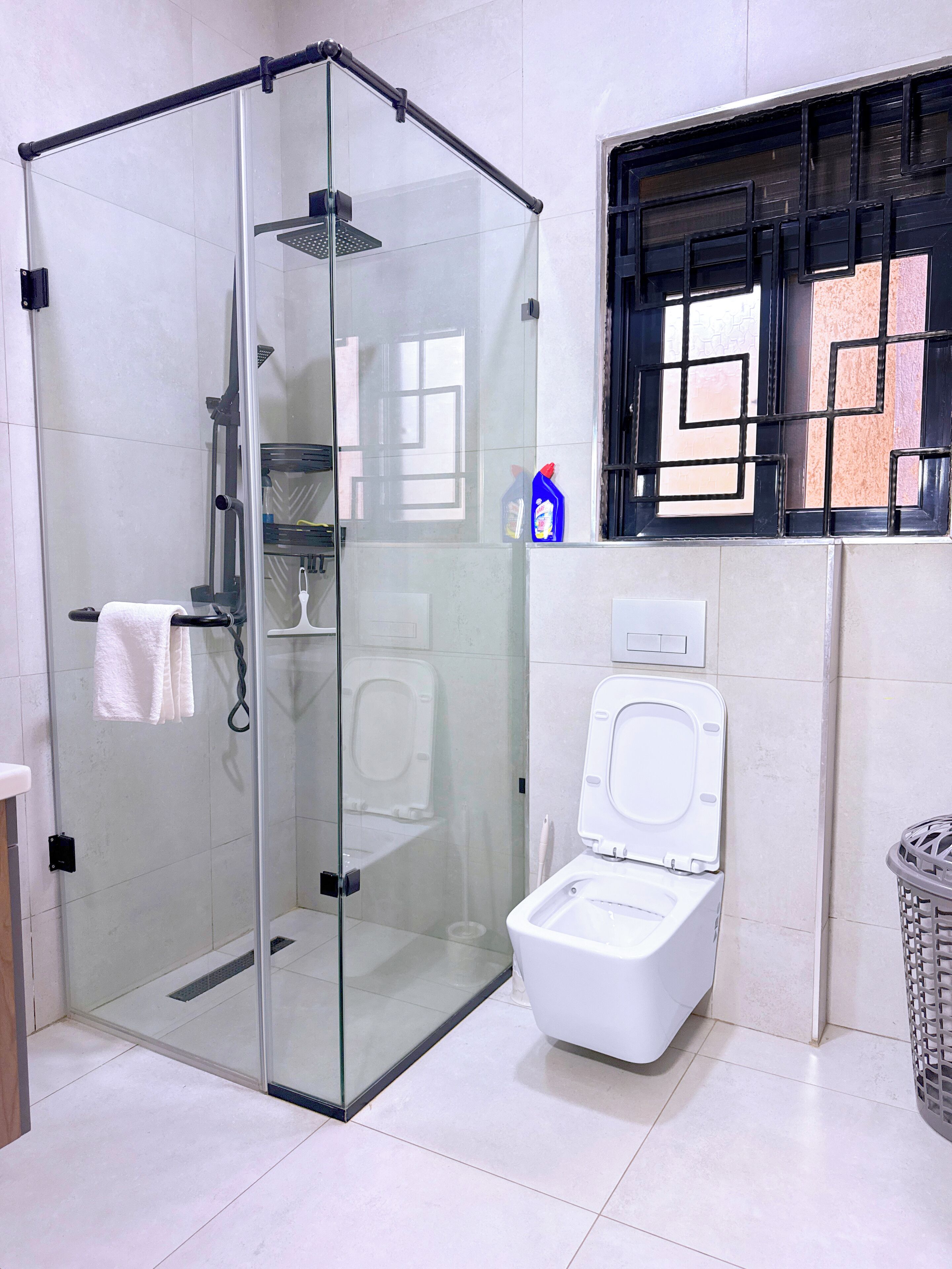 Un baño con ducha envuelta en vidrio, inodoro y ventana con diseño geométrico.