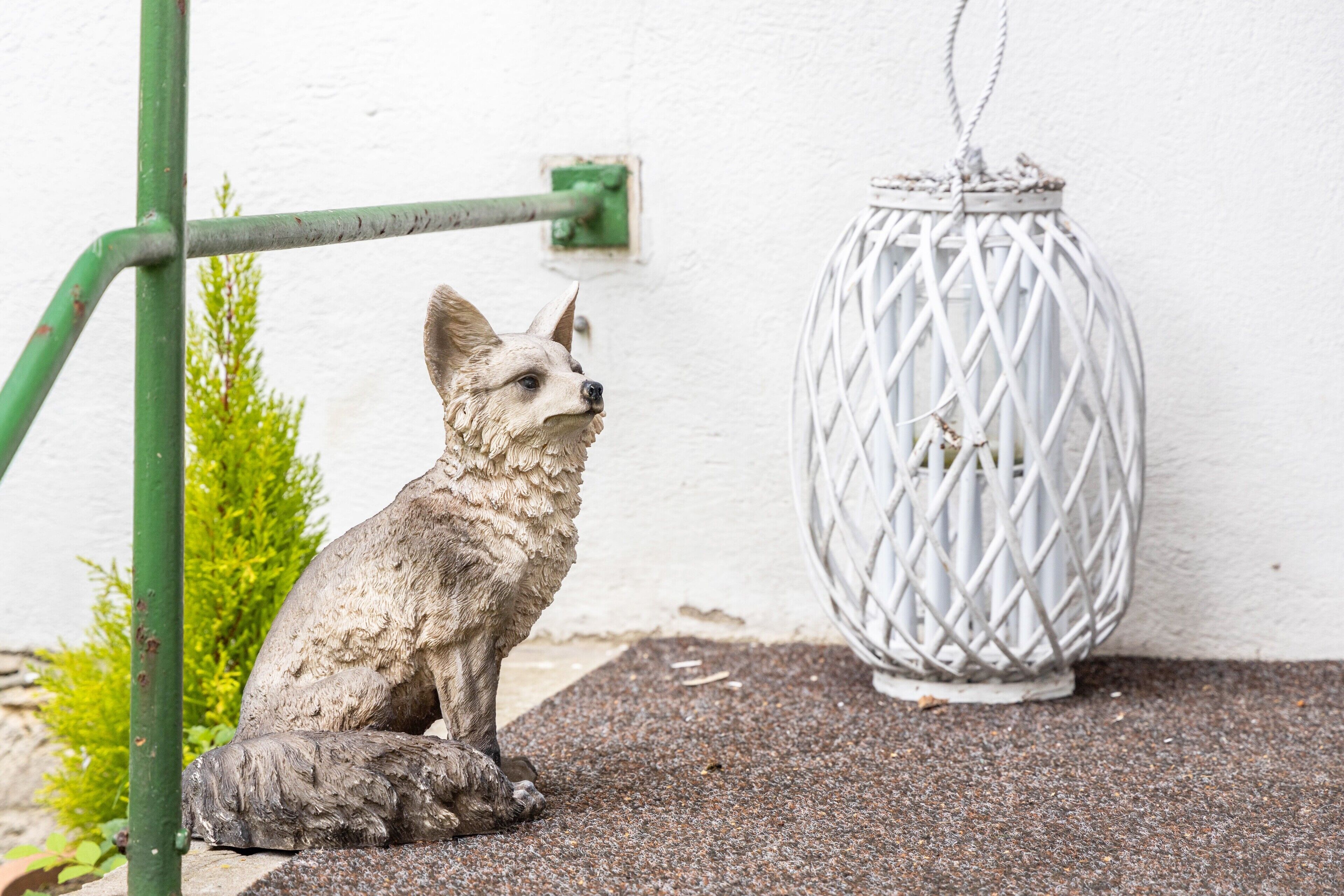 Eine Fox-Skulptur sitzt auf einer strukturierten Oberfläche in der Nähe einer weißen Wand und einer Metallkonstruktion.