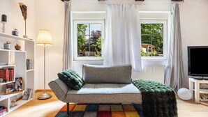 A cozy living room with a sofa, a colorful rug, a bookshelf, a lamp, and a view of the outdoors through a window.