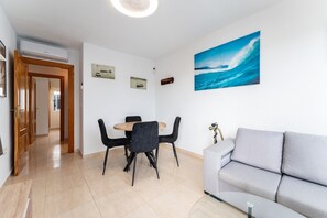 A modern living room with a grey sofa, a dining table with black chairs, a painting of a wave, and a ceiling fan.