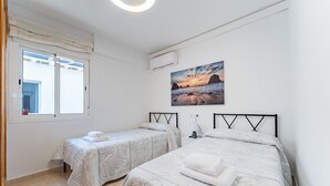 Une chambre avec deux lits, une fenêtre avec des rideaux, un tableau représentant une plage et un appareil de climatisation.