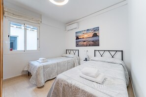 A bedroom with two beds, a window with curtains, a painting of a beach, and an air conditioning unit.