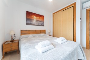 A bedroom with a bed, a nightstand, a wall-mounted lamp, a wooden wardrobe, and a sunset painting.