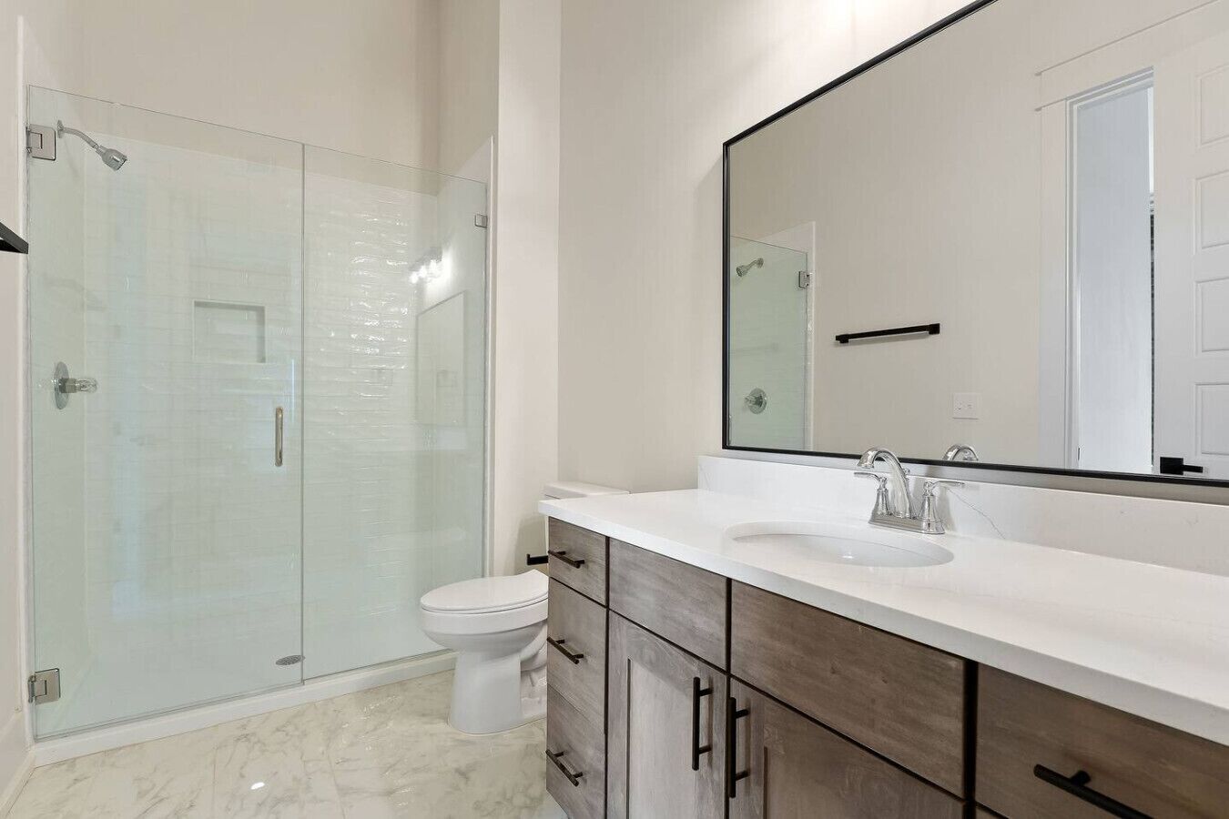 A modern bathroom with a glass shower enclosure, a white toilet, a large mirror, and a white sink with a wooden vanity.