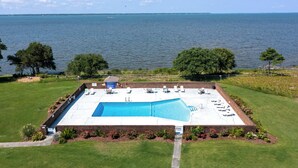 A rectangular swimming pool with a brick deck, surrounded by a grassy area and trees, with a view of the sea in the background. - The Sound of Family: Shared Pier | Community Pools and More! (Nags Head)