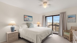 A bedroom with a bed, two bedside tables with lamps, a ceiling fan, a window with curtains, and a view of the beach. - The Sound of Family: Shared Pier | Community Pools and More! (Nags Head)