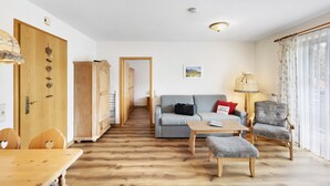 A living room with a sofa, armchair, ottoman, wooden dining table, and a hallway leading to another room.