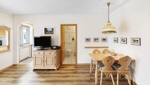 A room with a wooden cabinet, a television, a dining table with chairs, a hanging light fixture, and framed pictures on the wall.