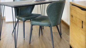 A dining area with a dark wooden table, two green upholstered chairs with metal legs, and a wooden cabinet to the right.