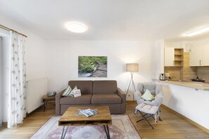 A modern living room with a sofa, a coffee table, and a chair. There is a kitchen area with cabinets and a countertop. A painting hangs on the wall.
