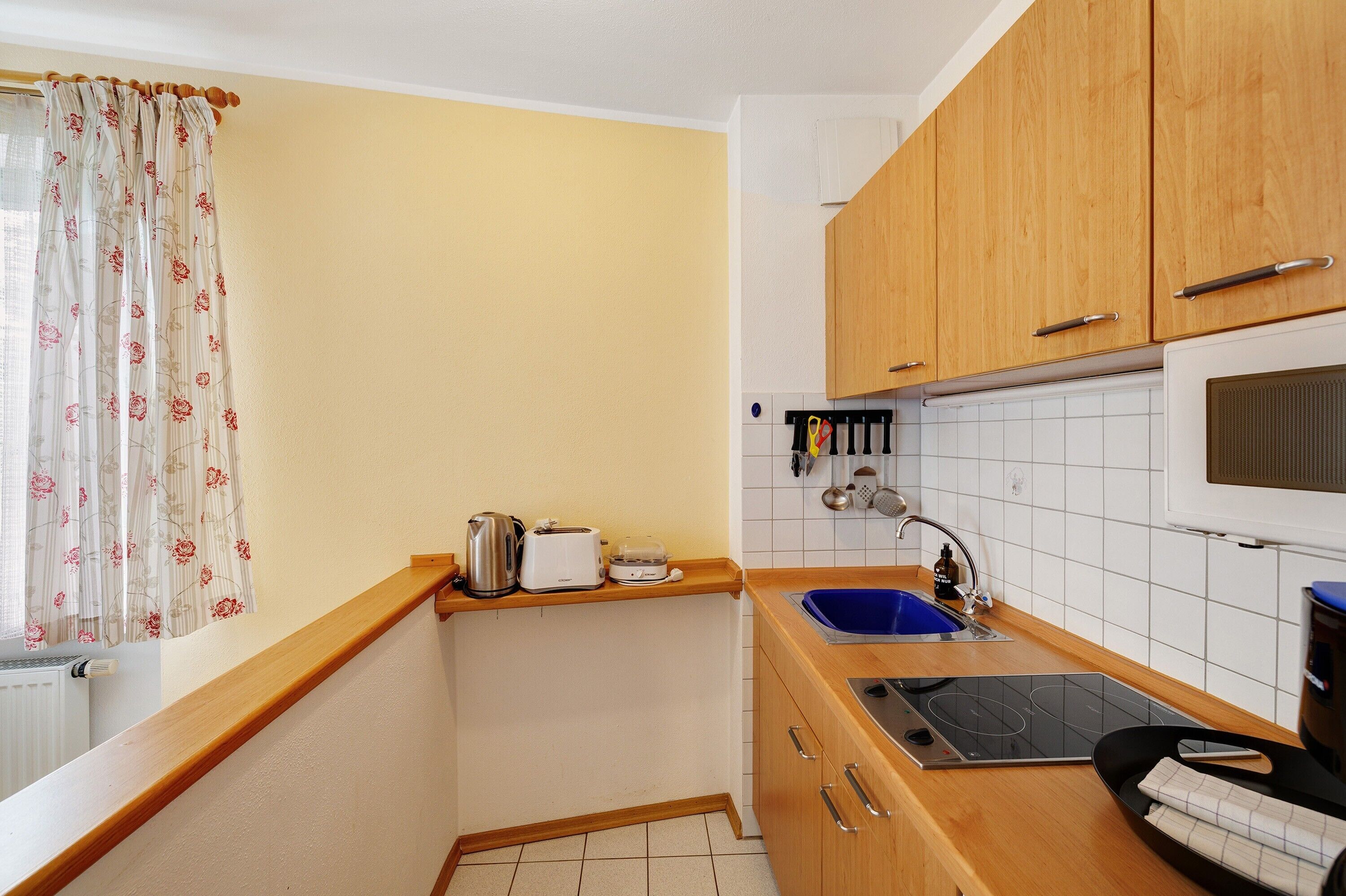 Cocina compacta con armarios de madera, estufa integrada, lavamanos y ventana con cortinas florales.