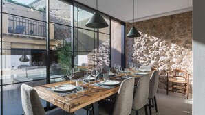 Comedor con una mesa de madera dispuesta para seis personas, rodeada de sillas grises con tapicería. Amplias ventanas ofrecen vista a un muro de piedra y vegetación exterior.
