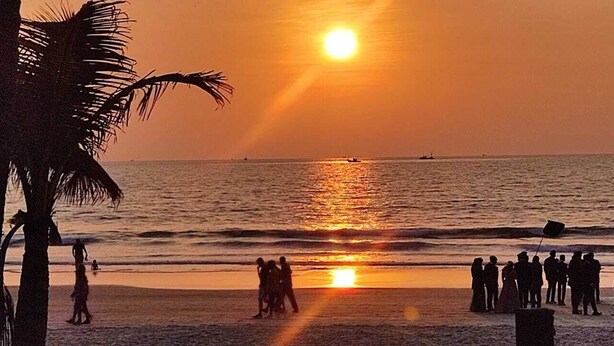 A beach scene with the sun setting over the ocean, palm trees, and silhouettes of people on the sand. - Furtados Beach Cottages  (Benaulim)