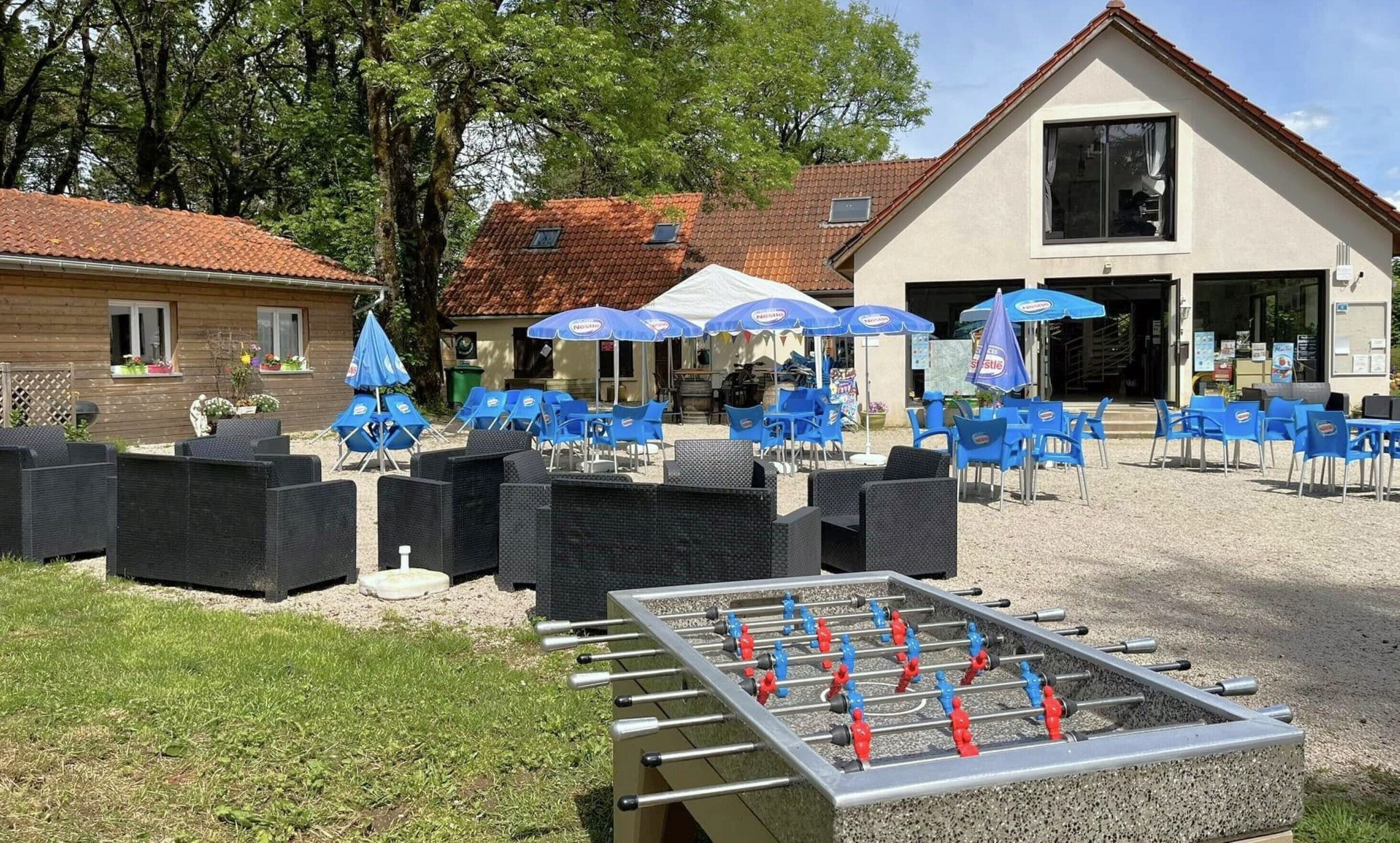 An outdoor seating area with blue chairs and umbrellas, a foosball table, and a building with a tiled roof in the background.