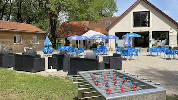 An outdoor seating area with blue chairs and umbrellas, a foosball table, and a building with a tiled roof in the background.