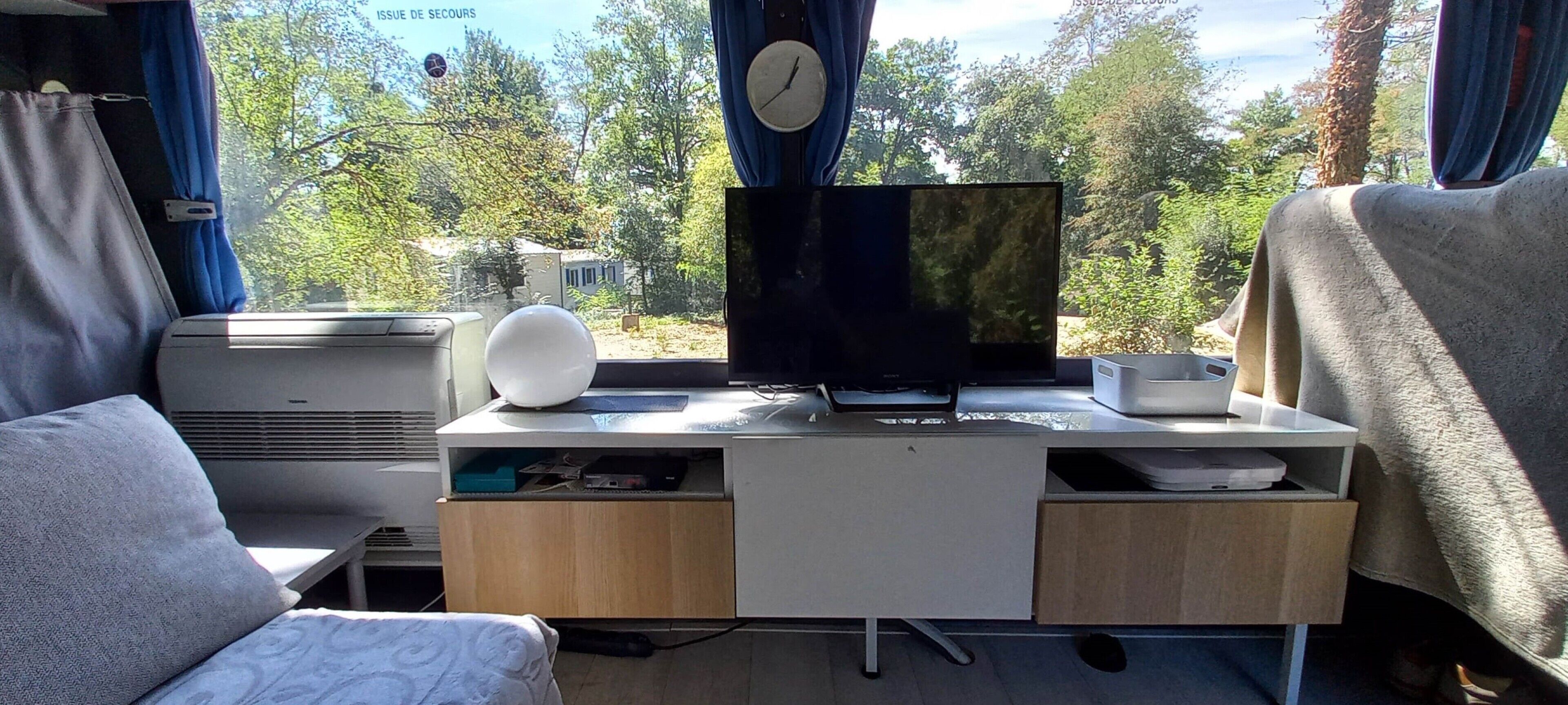 A compact living space with a bed, a desk with a computer monitor, a clock, and a cup.
