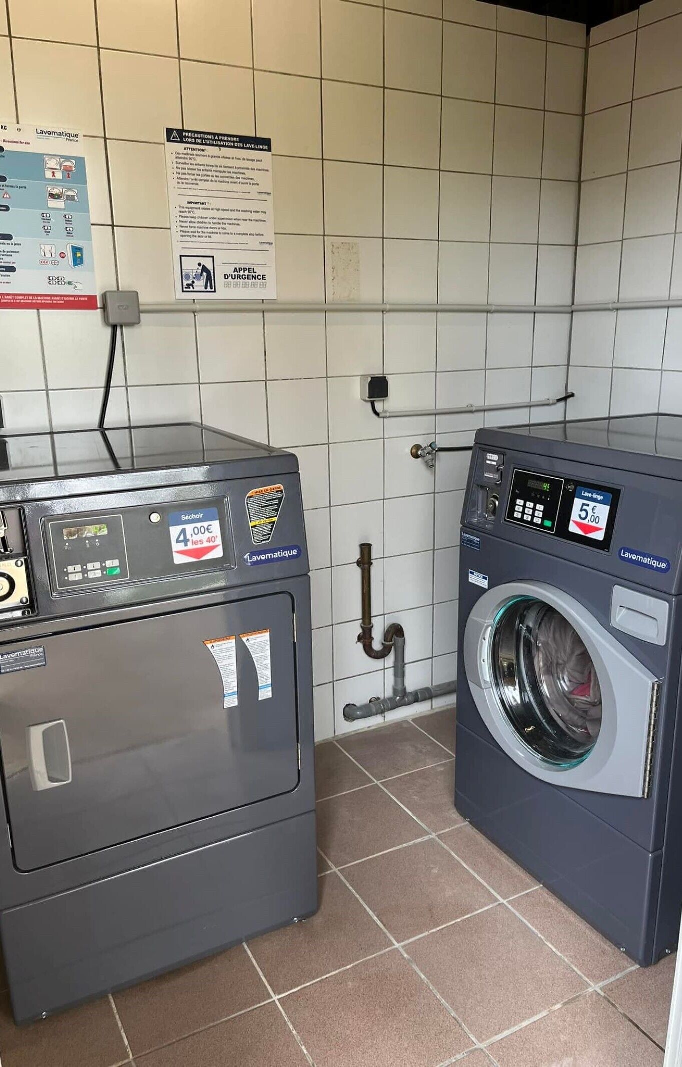Eine Wäscherei mit Waschmaschine und Trockner, einer Bedienkonsole und einem Aushang an der Wand.
