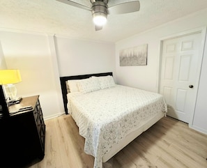 Un dormitorio con cama, ventilador de techo, mesita de noche con lámpara, una puerta y una obra de arte colgada en la pared.
