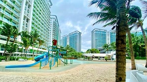 Zona de playa con un parque infantil, palmeras y edificios altos al fondo.