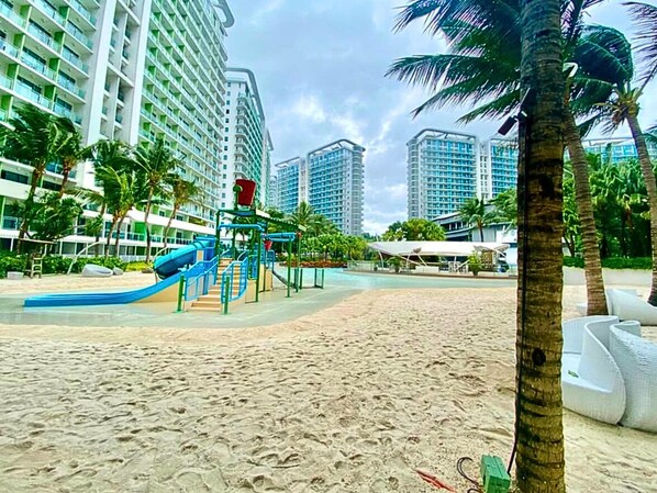 A beach area with a playground, palm trees, and high-rise buildings in the background.