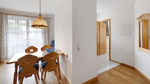 Uma área de refeições com mesa e cadeiras de madeira, um lustre pendurado e vista pela janela com cortinas.