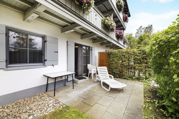 A patio with a table and two chairs, a wooden door, and a balcony with hanging flower baskets.