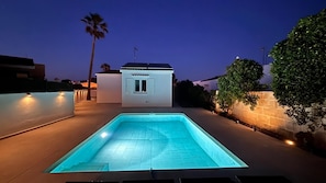 A swimming pool with a small building in the background, surrounded by palm trees and a stone wall.