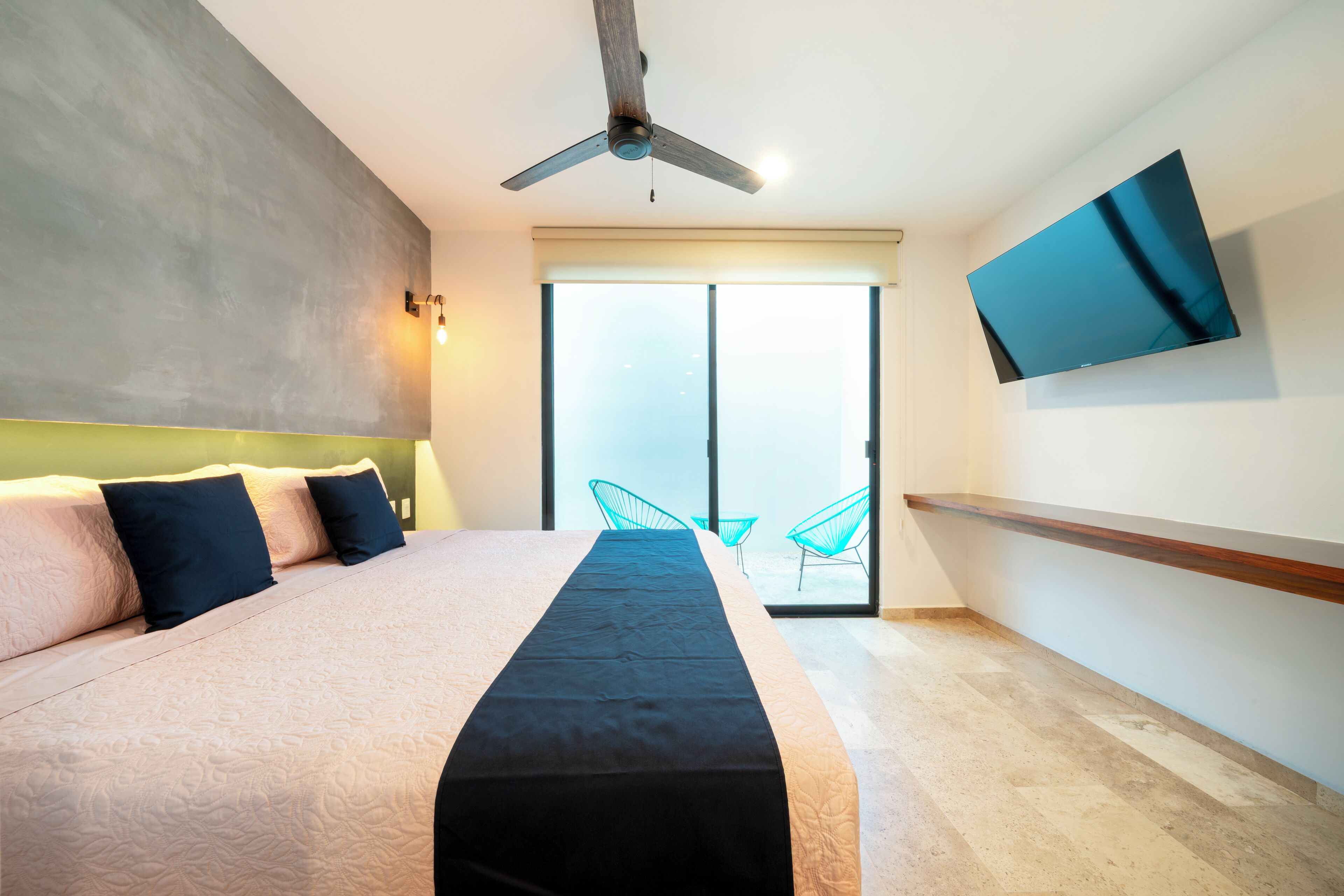 A bedroom with a bed, a ceiling fan, a TV mounted on the wall, and a sliding glass door leading to a balcony with chairs.