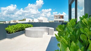 A rooftop terrace with a curved seating area, potted plants, and a view of the city skyline under a clear blue sky. - Comfortable studio with pool and gym - Huaya 101 (Playa del Carmen)