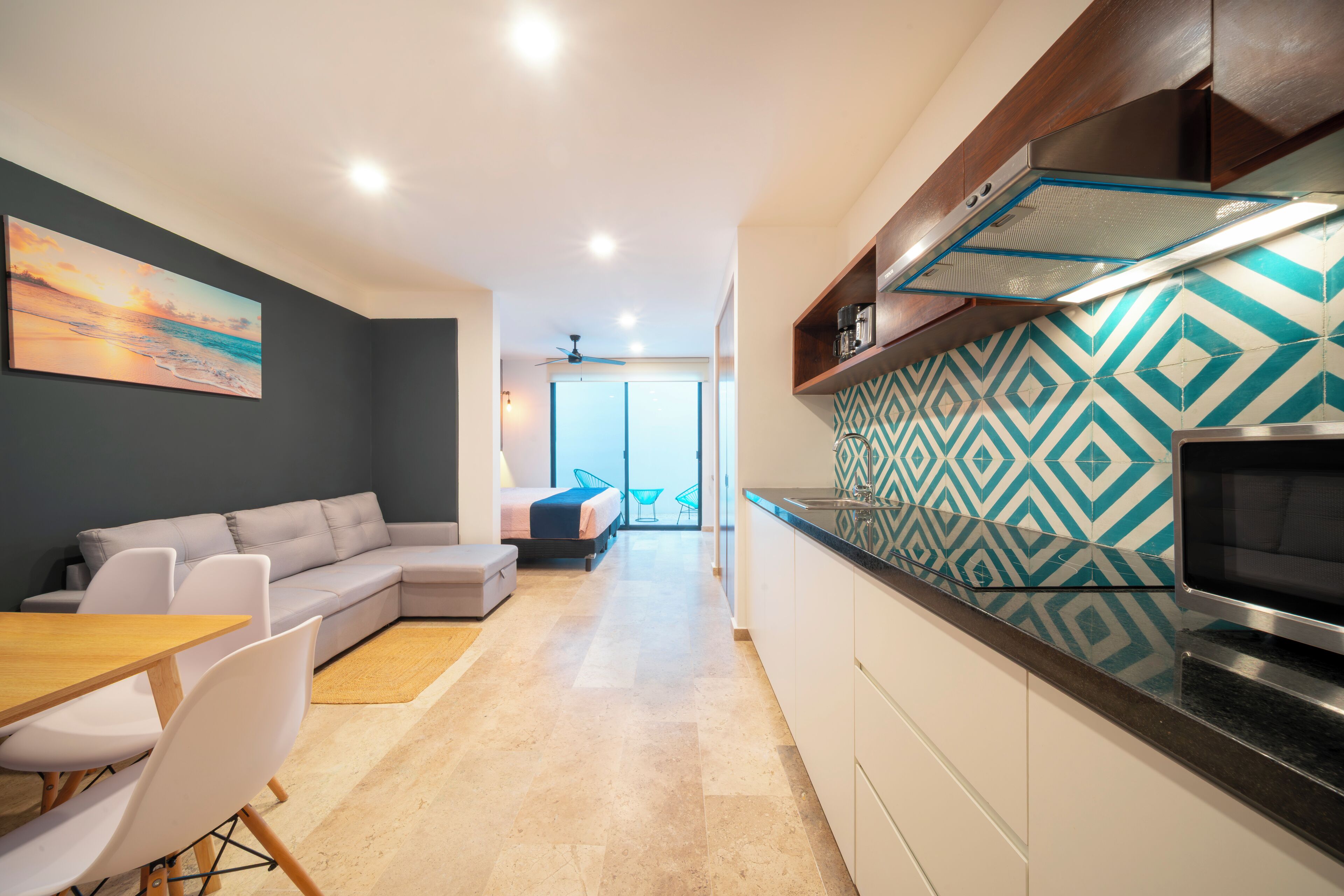 A modern kitchen with a patterned backsplash, a dining area with a wooden table and chairs, a gray sofa, and a view to a balcony.