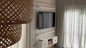 Een moderne woonkamer met een flatscreen televisie aan een witte televisiemeubel, een geweven mand die aan het plafond hangt, en dunne gordijnen voor de ramen.