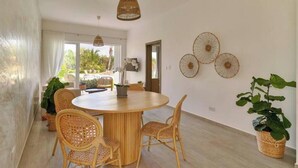 A spacious dining area with a round wooden table and wicker chairs, surrounded by large windows offering a view of greenery. - Garden View Relaxing Retreat (Punta Cana)